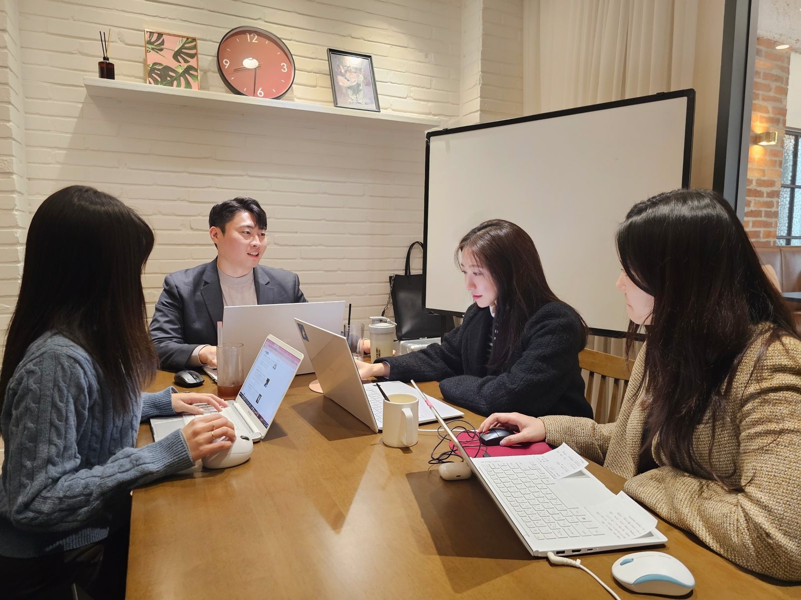 플라톤마케팅 회의실에서 네 사람이 긴 테이블에 마주 앉아 노트북과 인쇄물을 놓고 논의하는 모습. 뒤쪽에 화이트보드가 있다