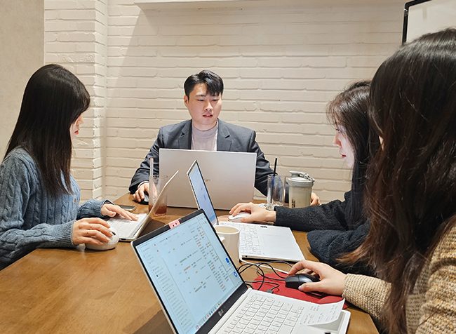 플라톤마케팅 구성원 네 명이 흰 벽돌 벽 회의실 테이블에 둘러앉아 각자 노트북 화면의 자료를 함께 보며 이야기하는 모습